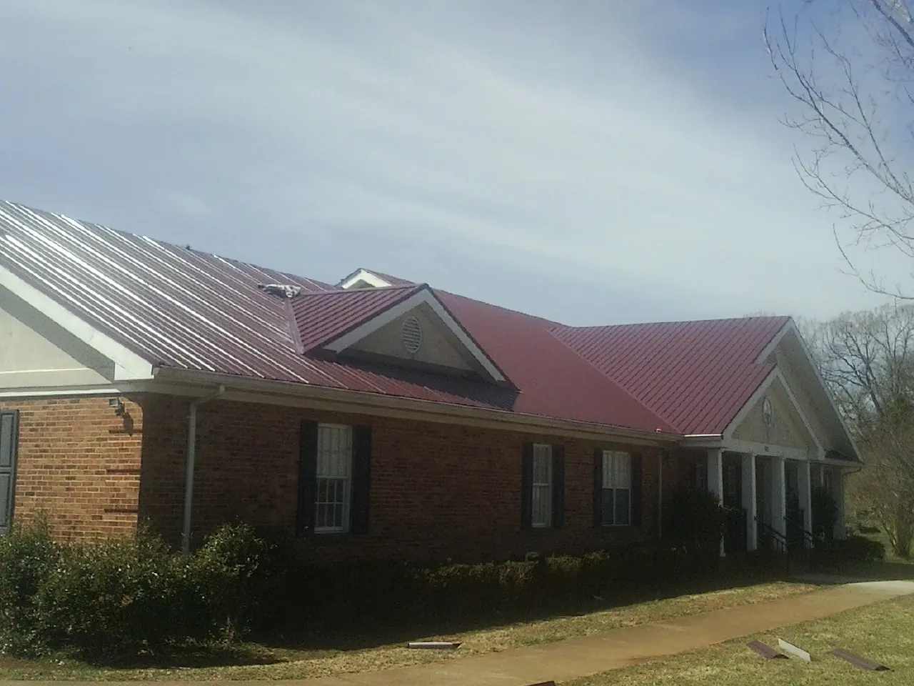 Red metal roof with gabled entries on a long brick building, from a Hail Damage Roof Repair job in Belgium