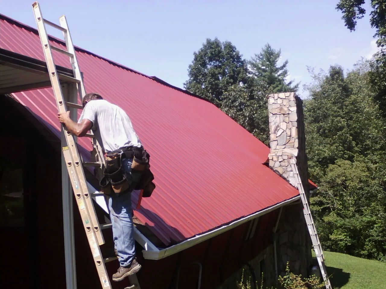 Roofer on a ladder beside a red metal roof and stone chimney — Soffit & Fascia Repair by Roofing Brothers in Belgium
